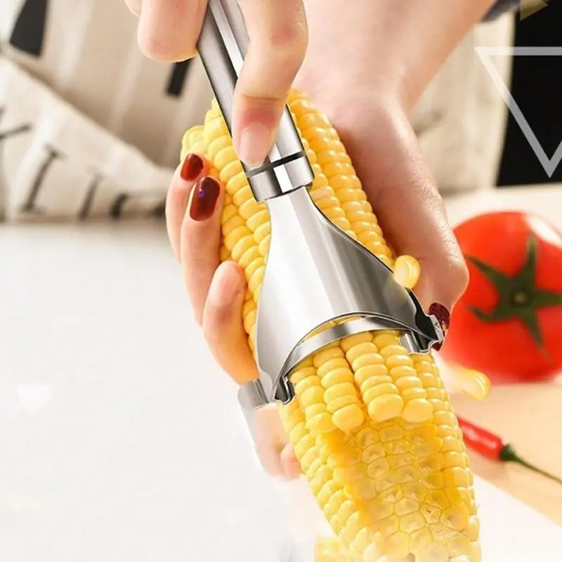 Hand using stainless steel corn peeler on corn cob in kitchen with tomato in background